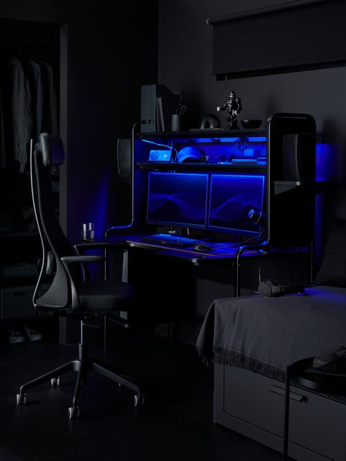 A black FREDDE desk is by a swivel chair in a dark, black bedroom. It holds two speakers, two monitors and blue LEDs inside.