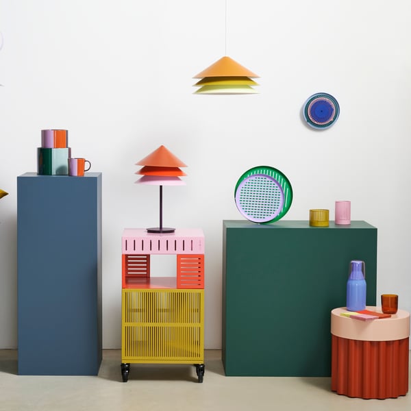 A TESAMMANS table lamp is on top of a storage unit on castors beside other colourful TESAMMANS items placed on pedestals.