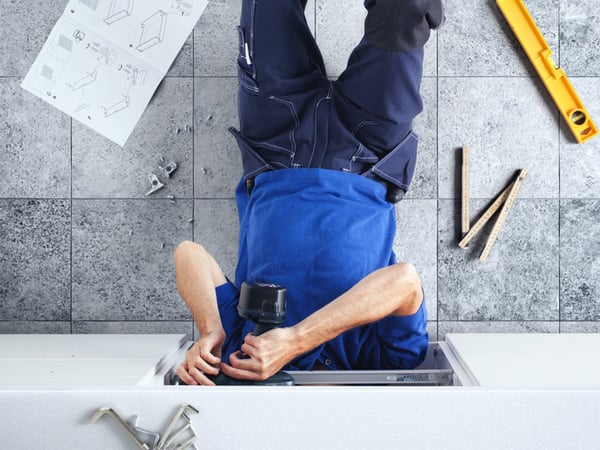 A person in blue work clothes is assembling furniture, head-down and using a power drill, with instructions and tools scattered around on a tiled floor.