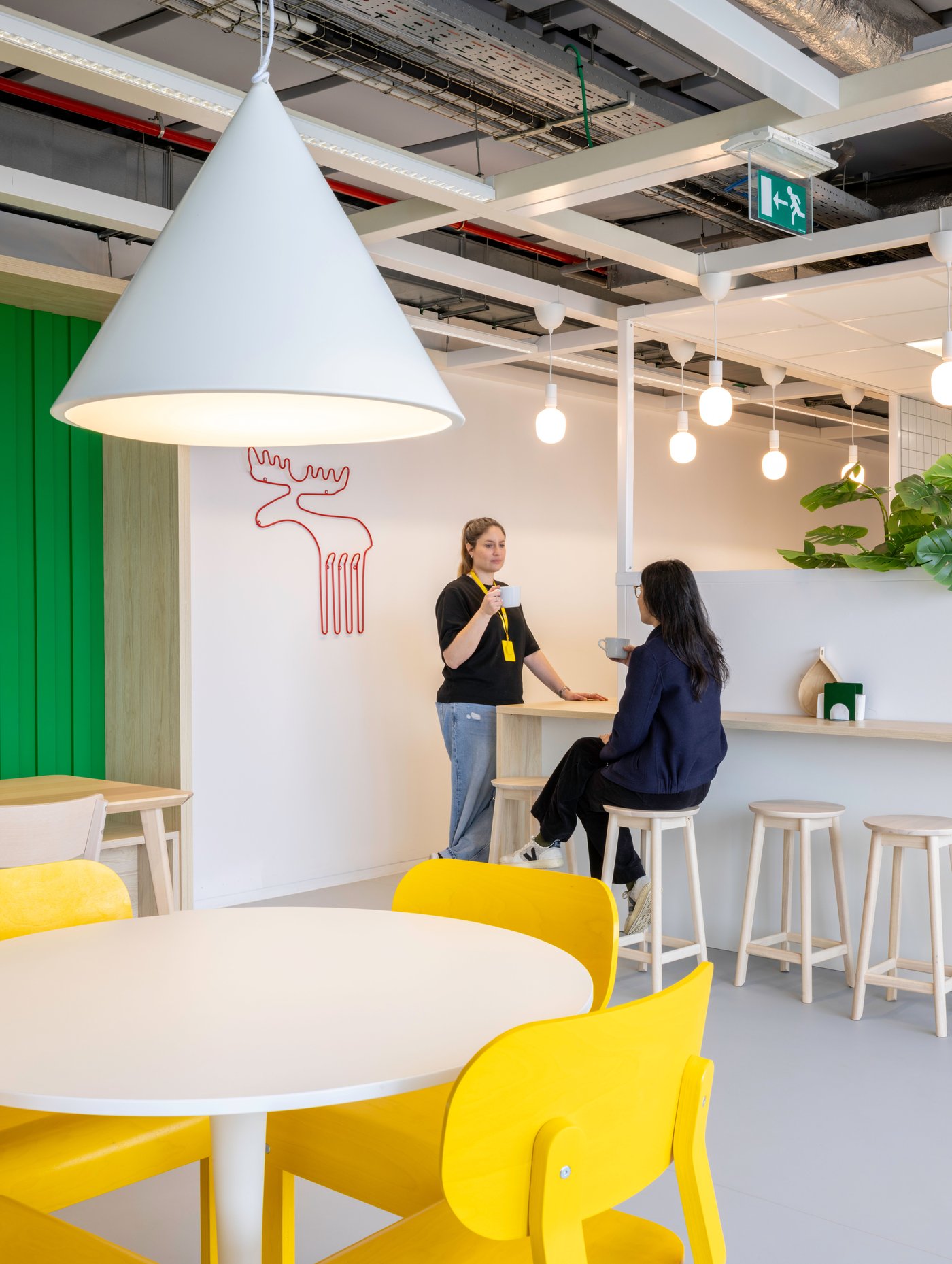 Un coin lumineux avec des assises IKEA jaunes, des tabourets en bois clair et un éclairage suspendu tout en douceur crée un espace accueillant pour discuter en toute décontraction et faire une pause-café rapide.
