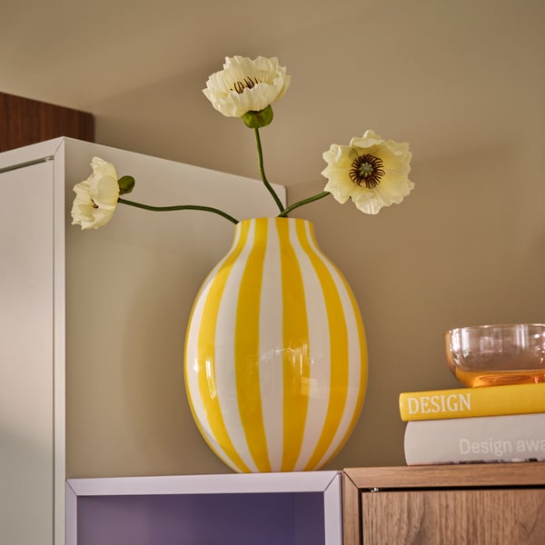 Ett vardagsrum med en vit- och gulrandig BLODBJÖRK vas fylld med blommor, placerad ovanpå en ljuslila EKET enhet.
