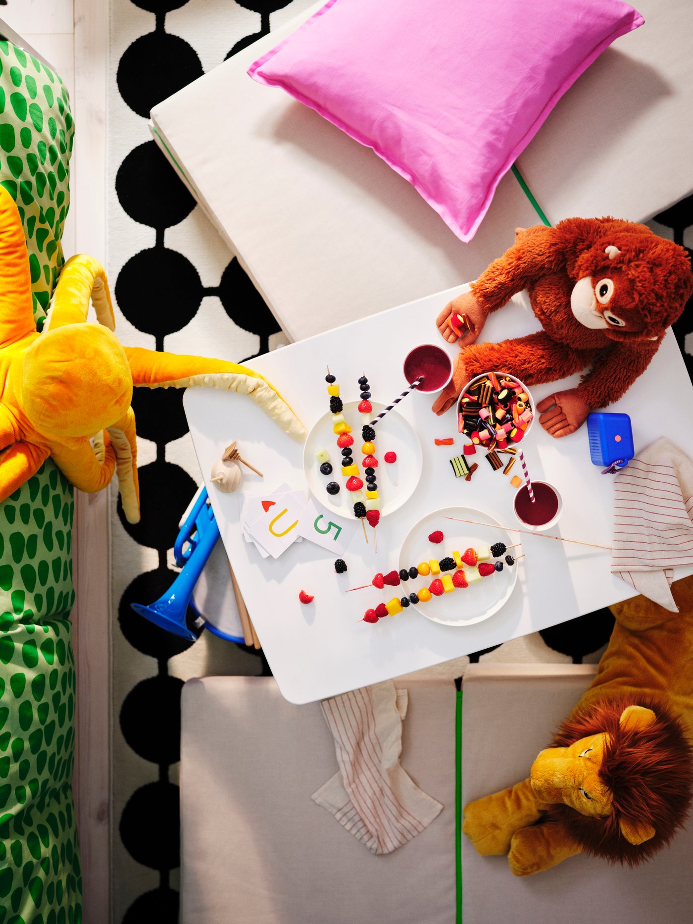 Picture taken from above a MAMMUT table set with playing cards, fruit, candy and soft toys. SLÄKT pouffe on floor around it.