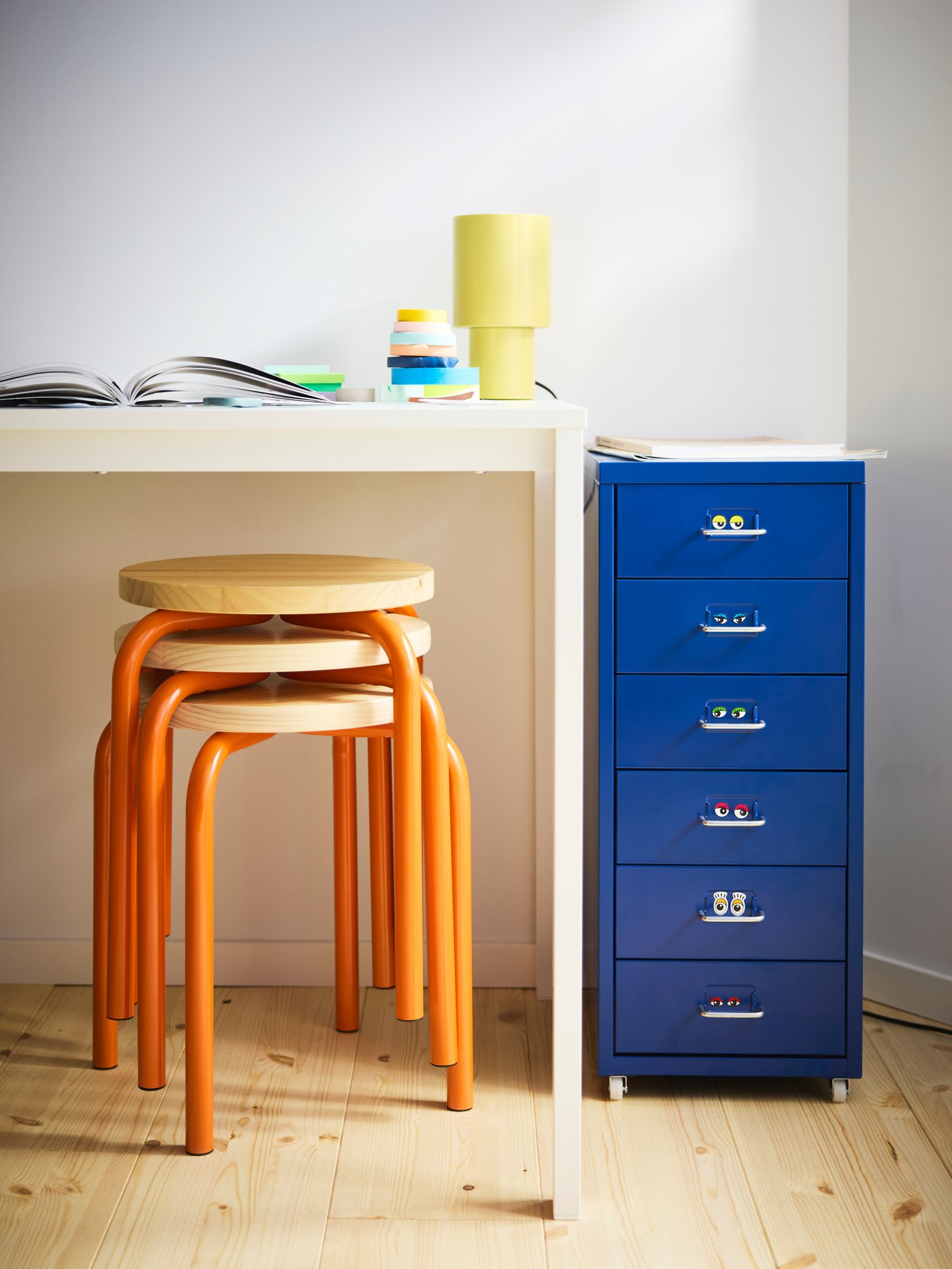 Närbild av en hög med orangea DOMSTEN stapelbara pallar under ett bord, och en blå hurts på hjul till höger.
