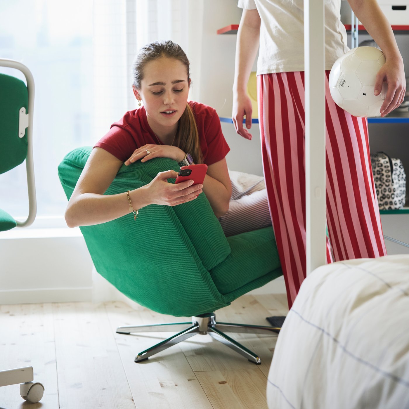 مراهق يجلس على كرسي DYVLINGE أخضر مستوحى من التصميم الكلاسيكي يتفاعل مع مراهق آخر يقف بجوار سرير.