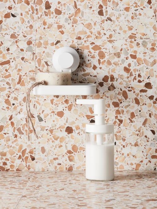 A glass TACKAN soap dispenser with a white plastic top sits on a terrazzo-style, tiled surface by a suction cup soap dish.