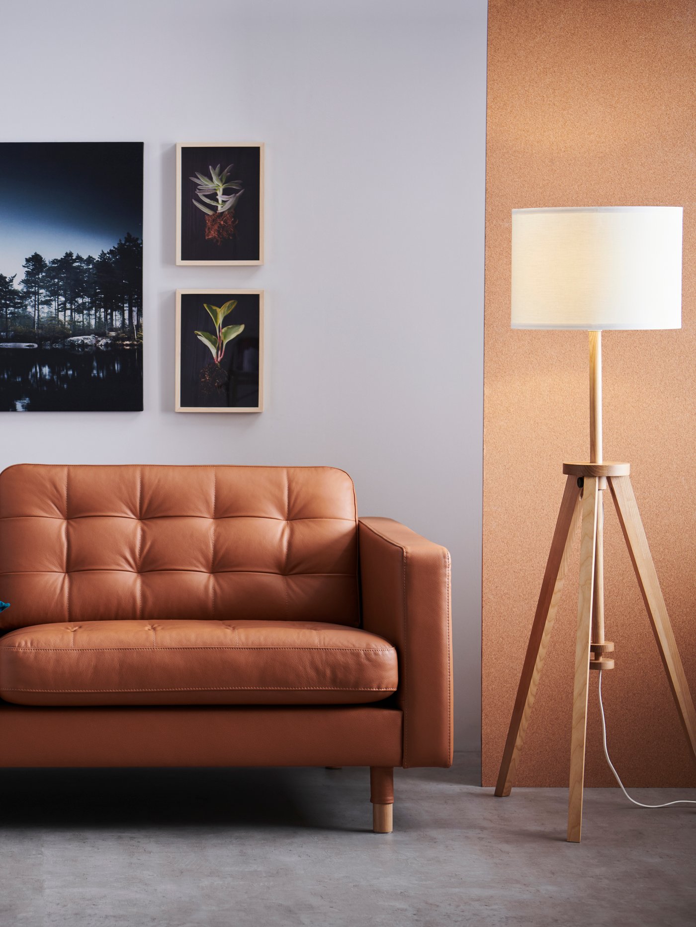 Salón con sofá de tres plazas con pantalla marrón dorado, lámpara de pie y tres cuadros colgados en una pared blanca.