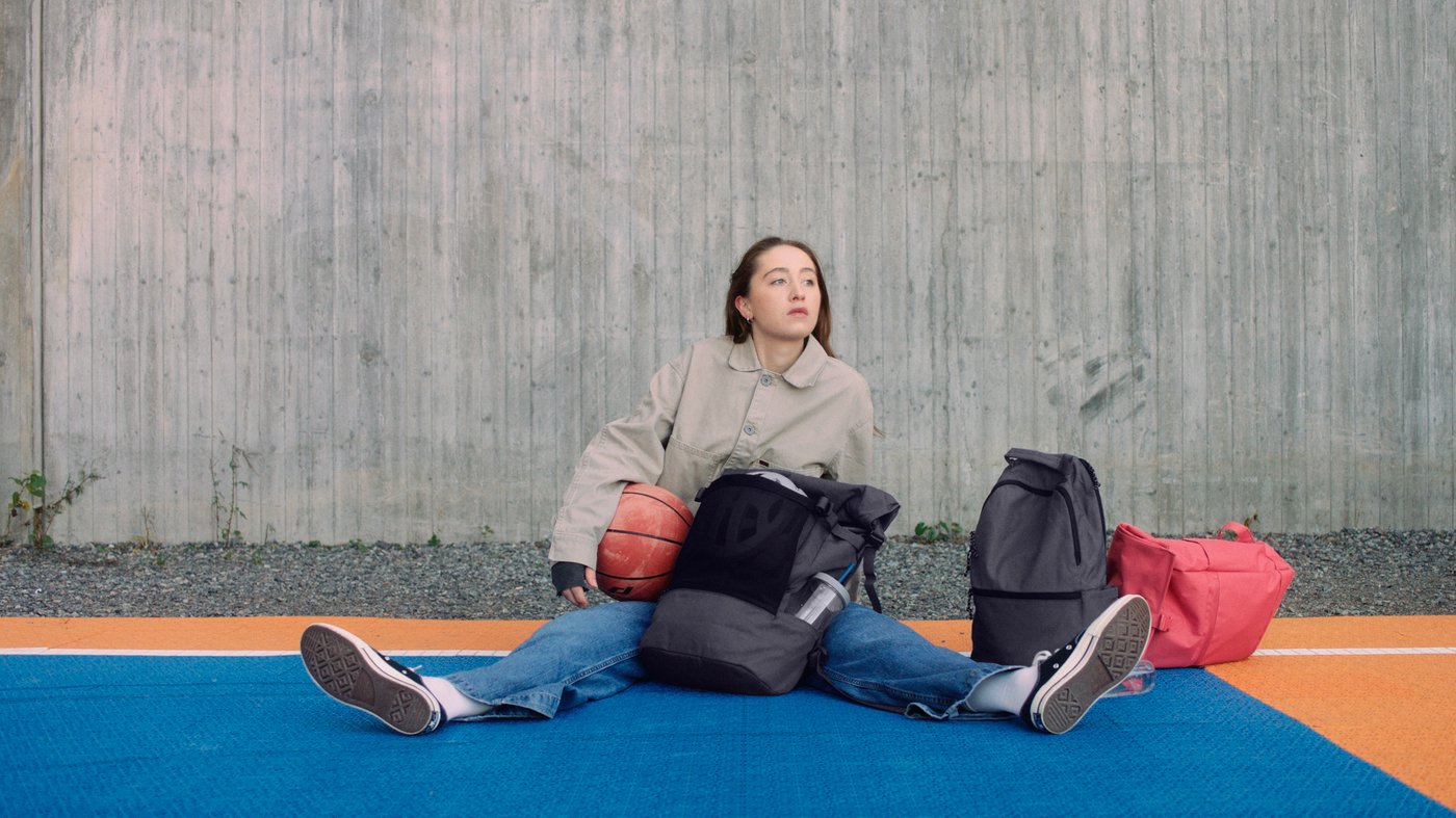 Ei jente sitter på en basketballbane med en basketball under armen og ryggsekkene DRÖMSÄCK, VÄRLDENS og STARTTID rundt seg.