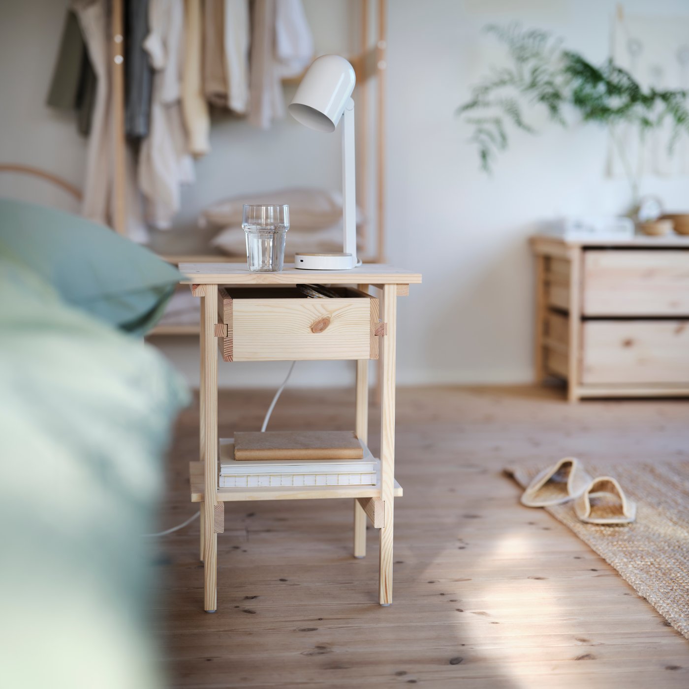 KRONÖREN sängbord i furu med låda och hylla som rymmer böcker, placerat bredvid en säng i ett ljust sovrum.