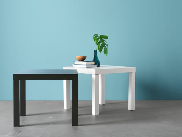 Dos mesas LACK, una negra y una blanca. La blanca tiene encima un par de libros y un florero con una planta verde.