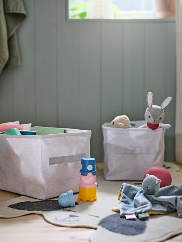 Two DRÖMSLOTT boxes on a floor holding a mix of toys, including a GULLIGAST squeaky soft toy. Toy are also on the floor.