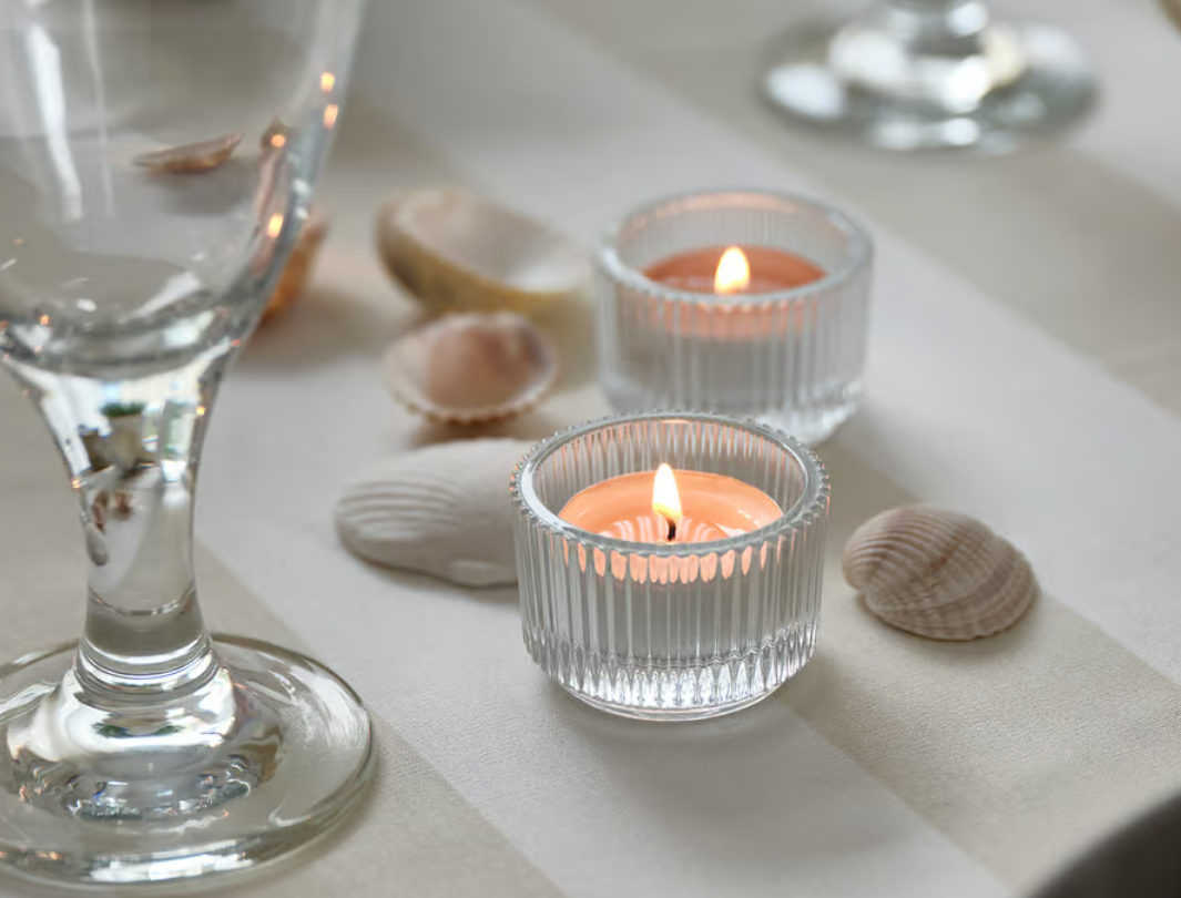 Teelichter in Glashaltern mit Muscheln und einem Weinglas auf hellem Tischläufer als dekorative Tischszene.