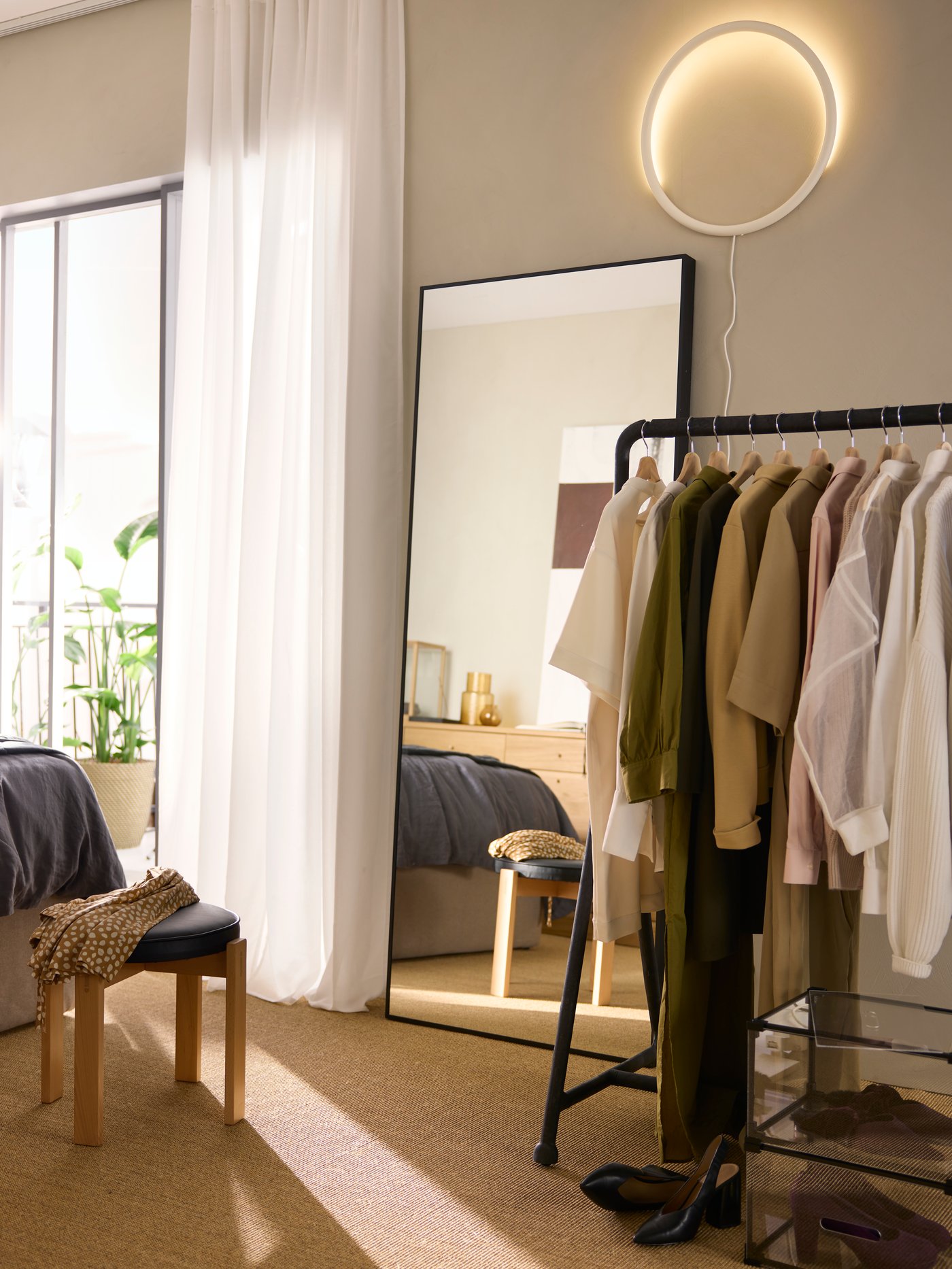 Ein schwarzer TURBO-Kleiderständer mit verschiedenen Kleidungsstücken in einem sonnigen, beigen Raum neben einem schwarz gerahmten HOVET-Spiegel.