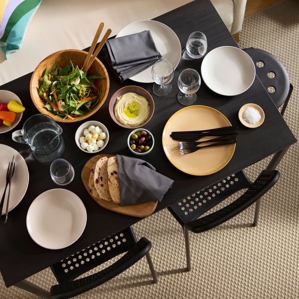 An aerial view of a black SANDSBERG table set with plates, SVALKA glasses, a salad, and a variety of foods served in bowls.