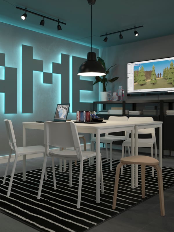 White TEODORES chairs around TOMMARYD tables placed on a GÖRLÖSE rug in a room with big, neon-backlit letters on the wall.