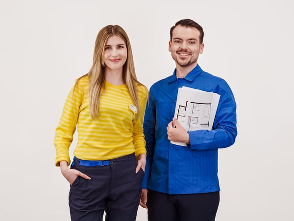 Projektant IKEA stojący w niebieskiej koszulce z projektami w ręku. Obok niego stoi konsultantka IKEA w żółtej koszulce.