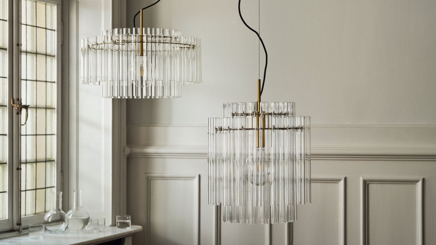 Two elegant, tiered glass tube pendant lamps with brass accents hang in a sophisticated room with light grey paneled walls and a large window.