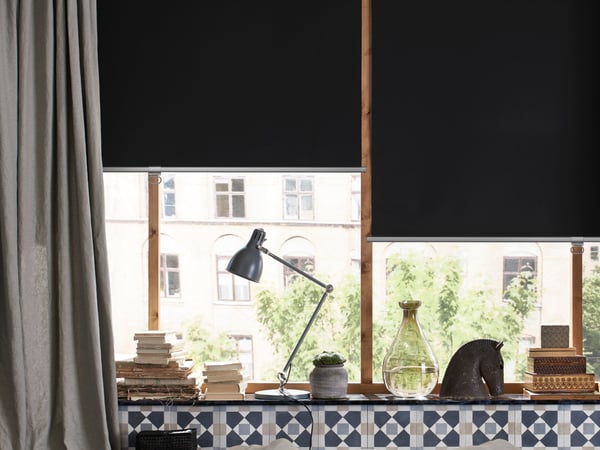 A well-lit interior space featuring a desk with a black adjustable desk lamp, stacks of books, and various decorative items. The desk is positioned in front of a large window with partially drawn black blinds, offering a view of green trees and the facade of an opposite building.