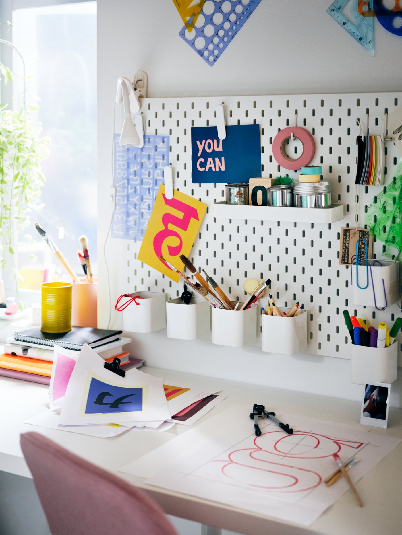 SKÅDIS blanco lleno de accesorios de colores, sobre una mesa LAGKAPTEN blanca y una silla giratoria LÅNGFJÄLL rosa.