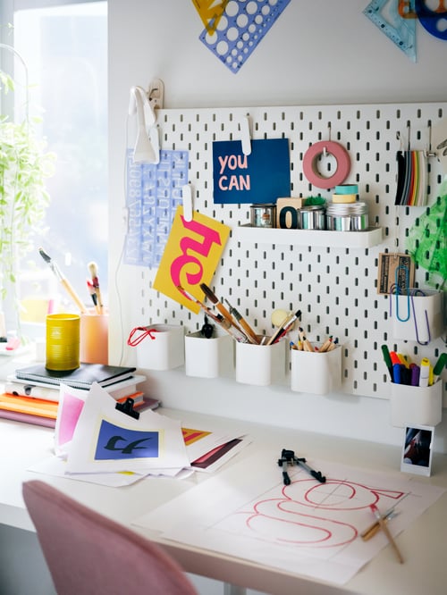 A white SKÅDIS pegboard filled with colourful accessories, over a white LAGKAPTEN tabletop and pink LÅNGFJÄLL swivel chair.