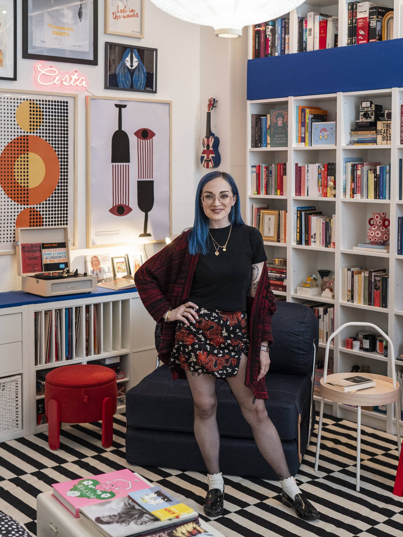 La content creator Teresa Agostini in piedi vicino a una poltrona blu jeans in un soggiorno colorato, con tappeto a righe, librerie piene di libri e poster grafici alle pareti.