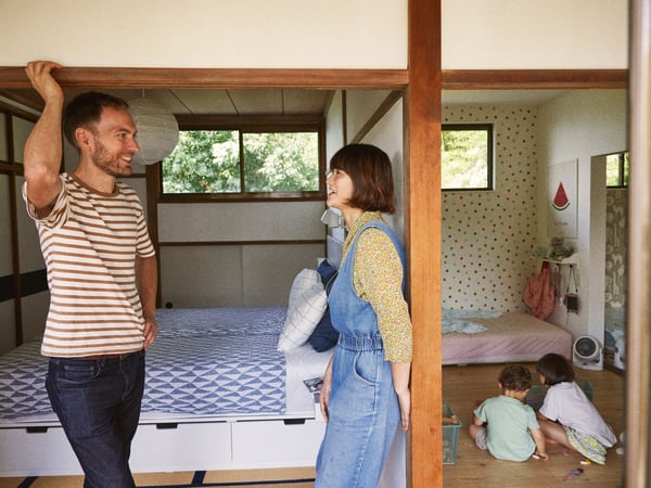 Una mujer y un hombre sonriendo dialogando entre sí dentro de una habitación con niños jugando en el suelo.