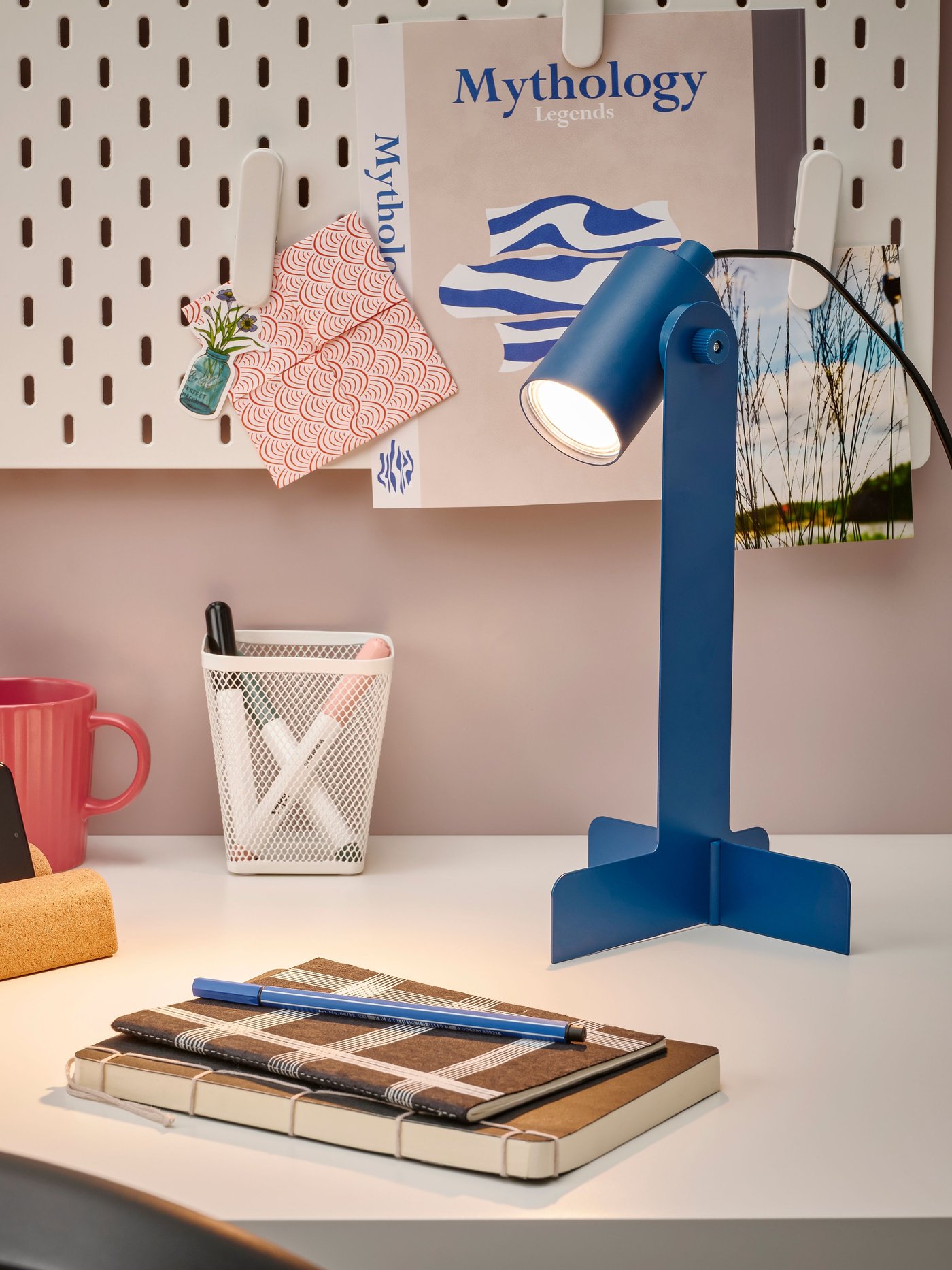 A dark blue FLOTTILJ desk lamp in front of a white SKÅDIS pegboard and next to a mug, notebooks and pens on a white desk.