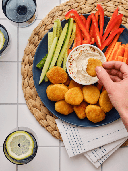 A snack plate with various vegetables, crispy wheat pieces, and a dip on a blue plate on a placemat.