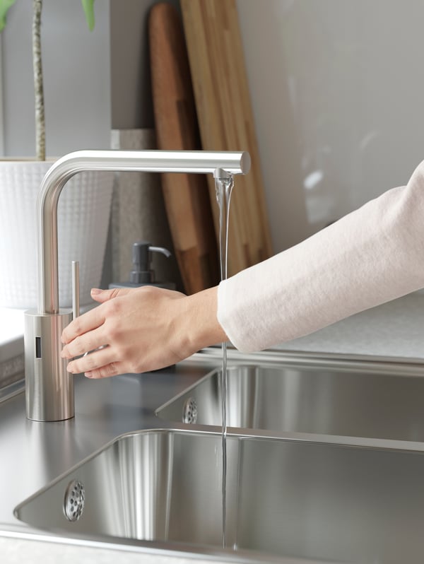 Eine Person mit einem weißen Oberteil aktiviert die IKEA TÄMNAREN Mischbatterie mit Sensor in Edelstahl und schaltet so das Wasser ein.
