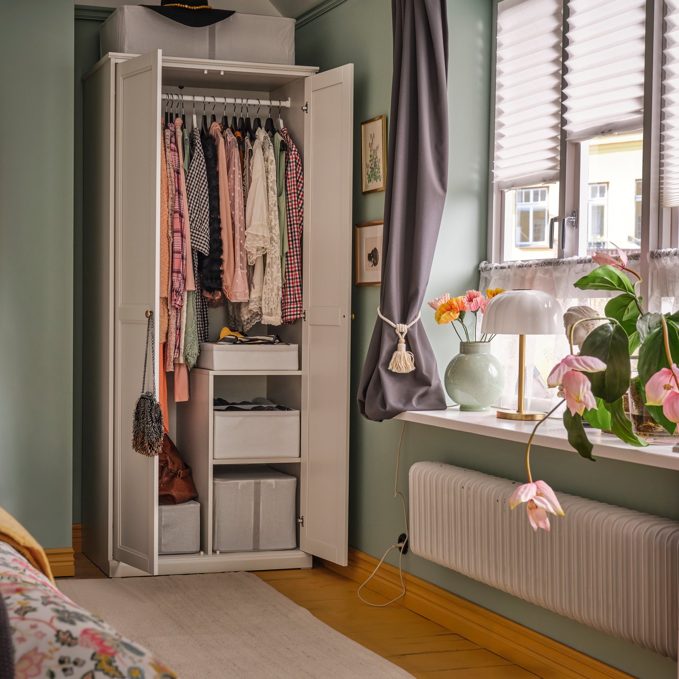 Camera da letto con guardaroba FÅGELFJÄLLET bianco sporco, con abiti e scatole contenitore all’interno, accanto a una finestra con piante.