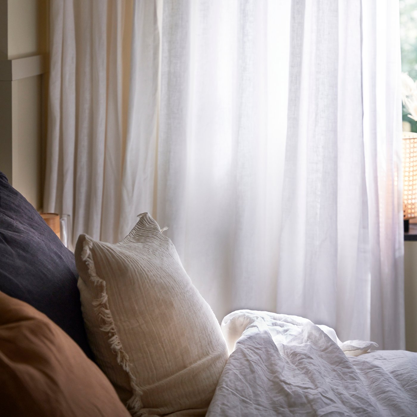 Närbild av en bäddad säng med mjuka kuddar i neutrala färger och vita skira gardiner som släpper in mjukt dagsljus från fönstret.
