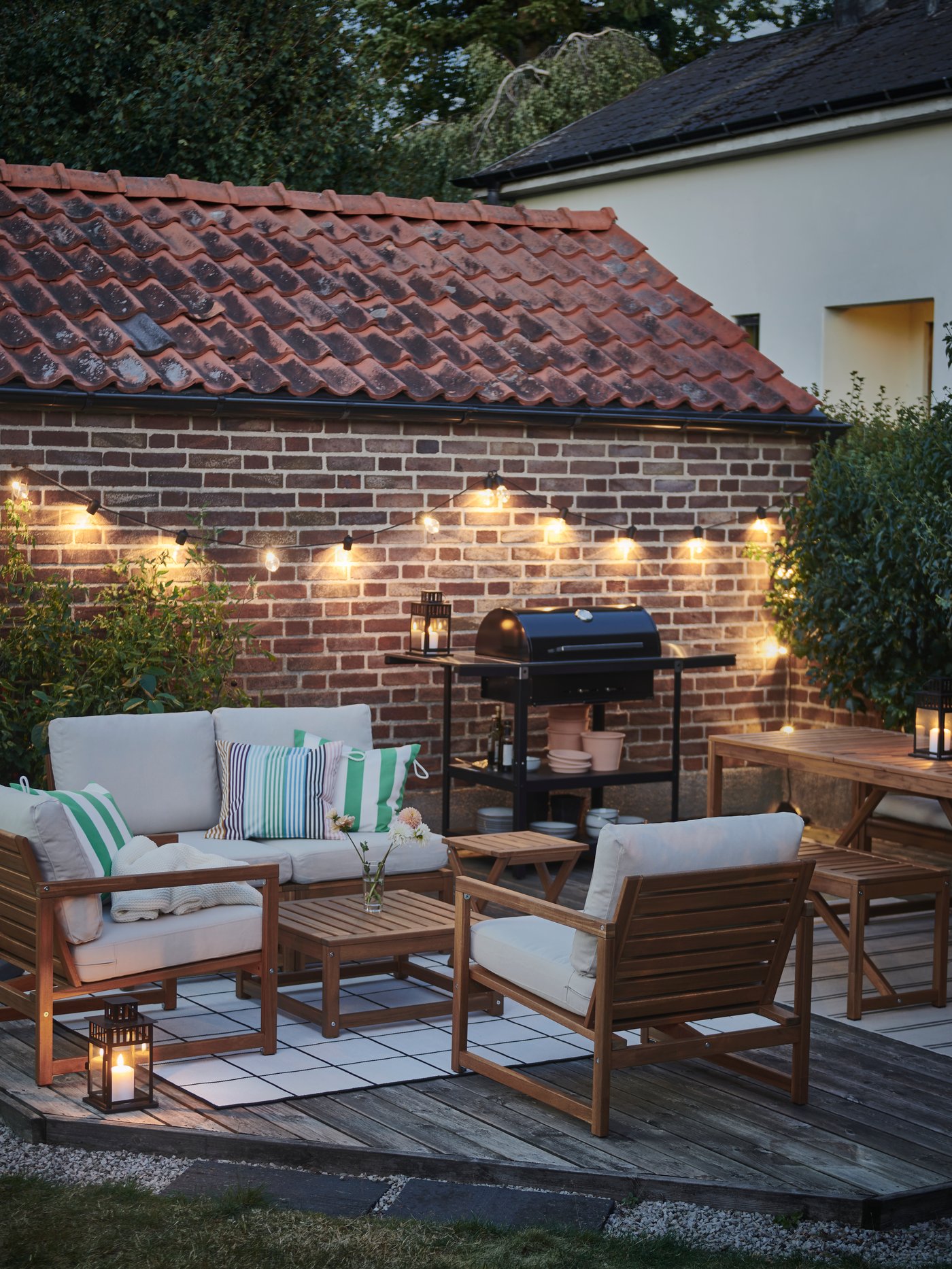 Es ist ein NÄMMARÖ Tisch mit 4 Stühlen in der Dämmerung zu sehen, beleuchtet von einer SVARTRÅ LED-Lichterkette. An der Wand eines Schuppens steht ein GRILLSKÄR Holzkohlegrill.