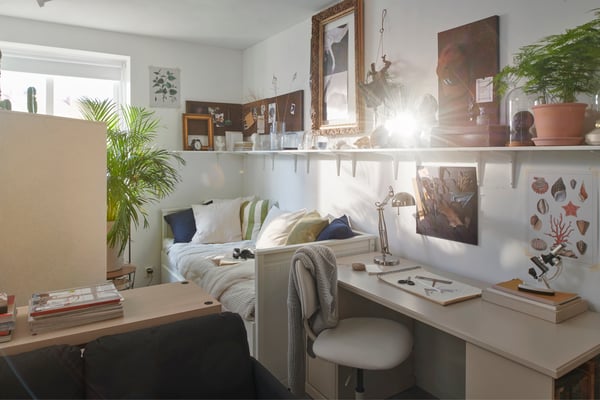 Chambre d’étudiant très éclairée. Lit d’appoint HEMNES dans le coin d’une pièce, des étagères murales avec divers objets. Bureau beige et chaise à côté du lit.