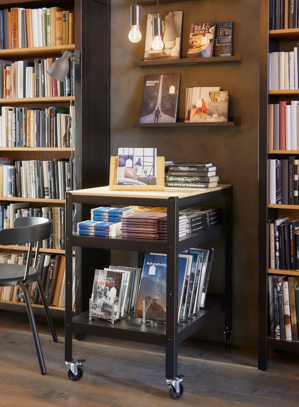 Wózek na kółkach z dwoma półkami wypełnionymi książkami, ustawiony pod brązową ścianą w wolnej przestrzeni między dwoma dużymi regałami.
