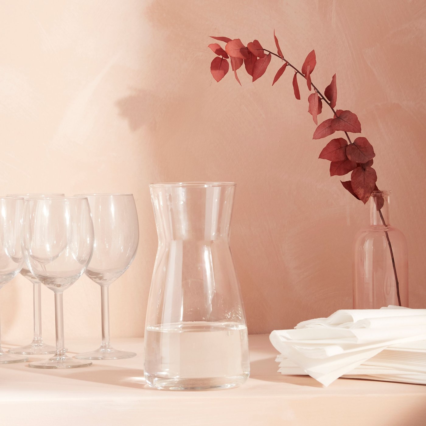 Three IKEA SVALKA wine glasses with a matching glass jug on a pink background, white napkins on the side.