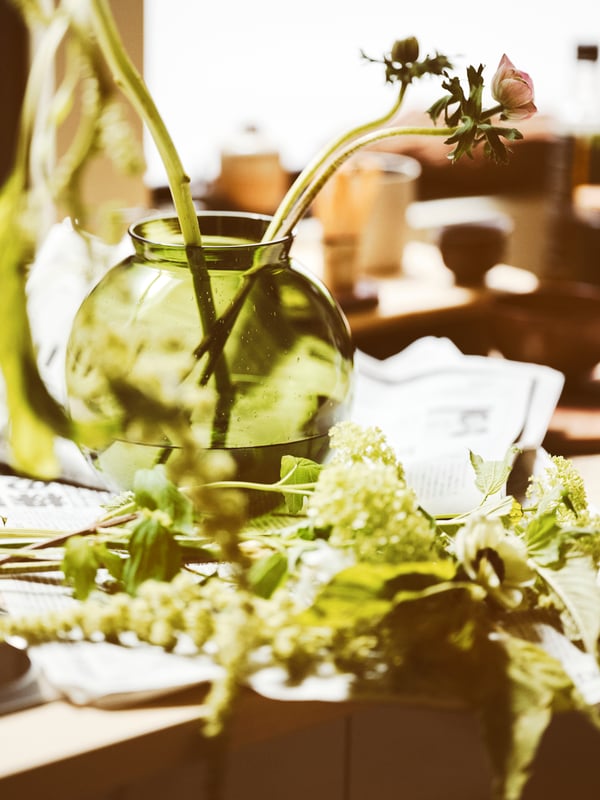 A green KONSTFULL vase with water and several stemmed flowers set on a surface along with other green plants and flowers.