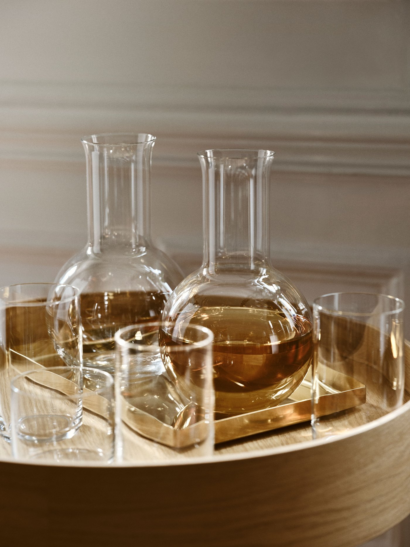 Two STOCKHOLM 2025 glass carafes stand on a brass-colored tray next to various glasses on a side table with oak veneer.