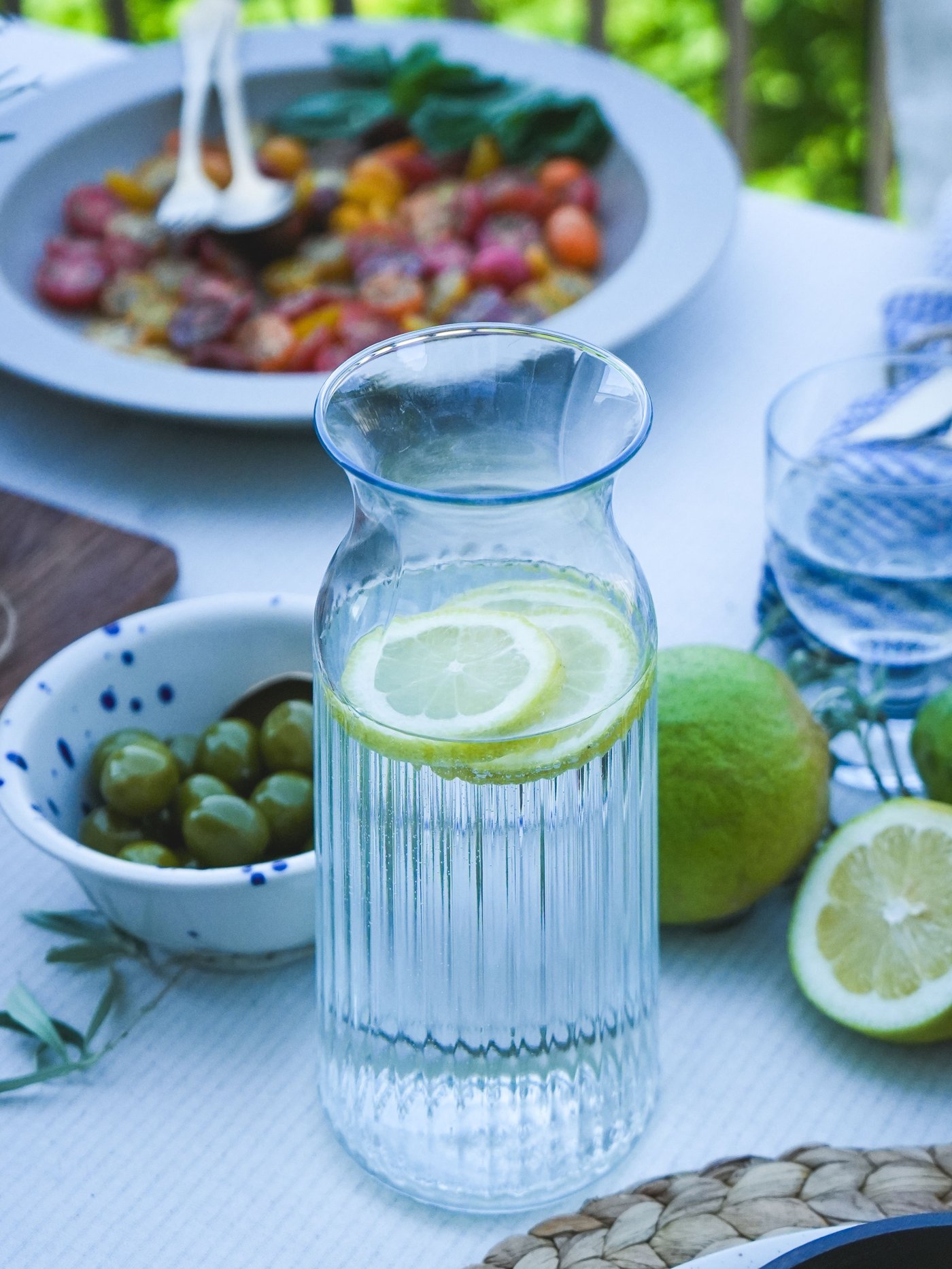 Décoration de table avec une carafe VARDAGEN remplie d'eau citronnée, des olives vertes et une salade de tomates au basilic, le tout entouré de citrons verts et de citrons frais.