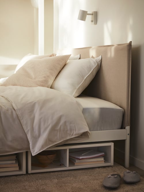 The top end of a KLEPPSTAD bed covered in ÄNGSLILJA bed linen, with a lamp on the wall above and a pair of slippers on the floor.