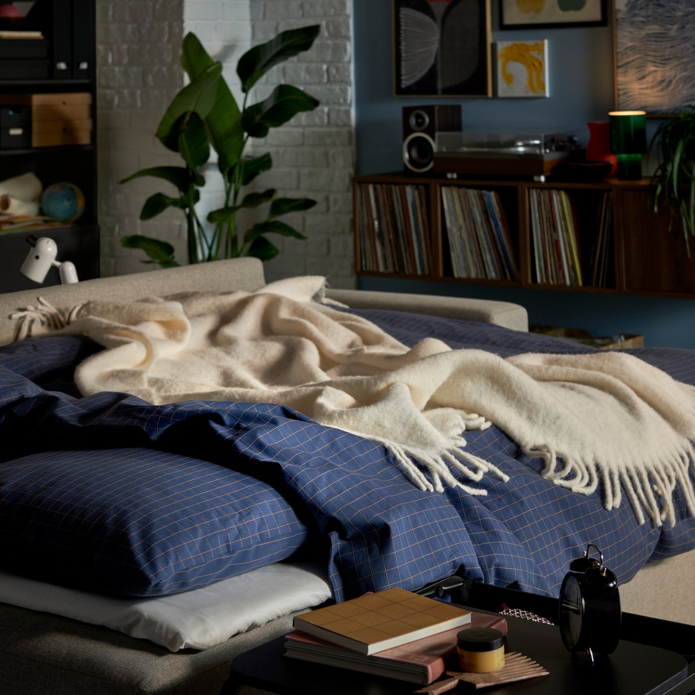 A cosy bed with blue-checked bedding, a white throw and a green, leafy potted plant in a bright bedroom setting.