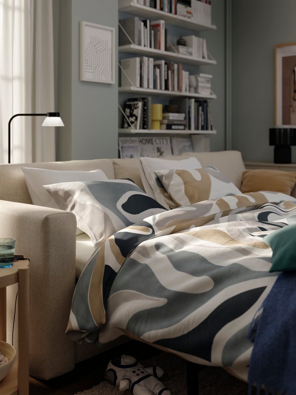 A beige FINNALA sofa-bed, shown as a bed, with multicolour MYGGLASVINGE bed linen, stands near a window.