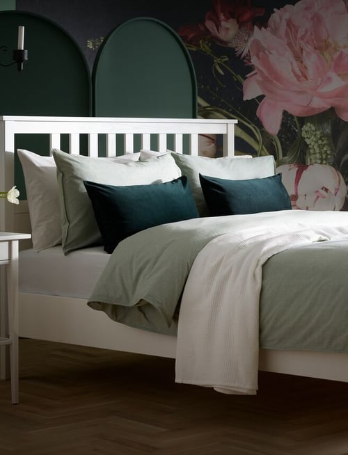 Bedroom with floral wallpaper and white solid pine bed frame with green bed linens, cushions, and white bedspread.