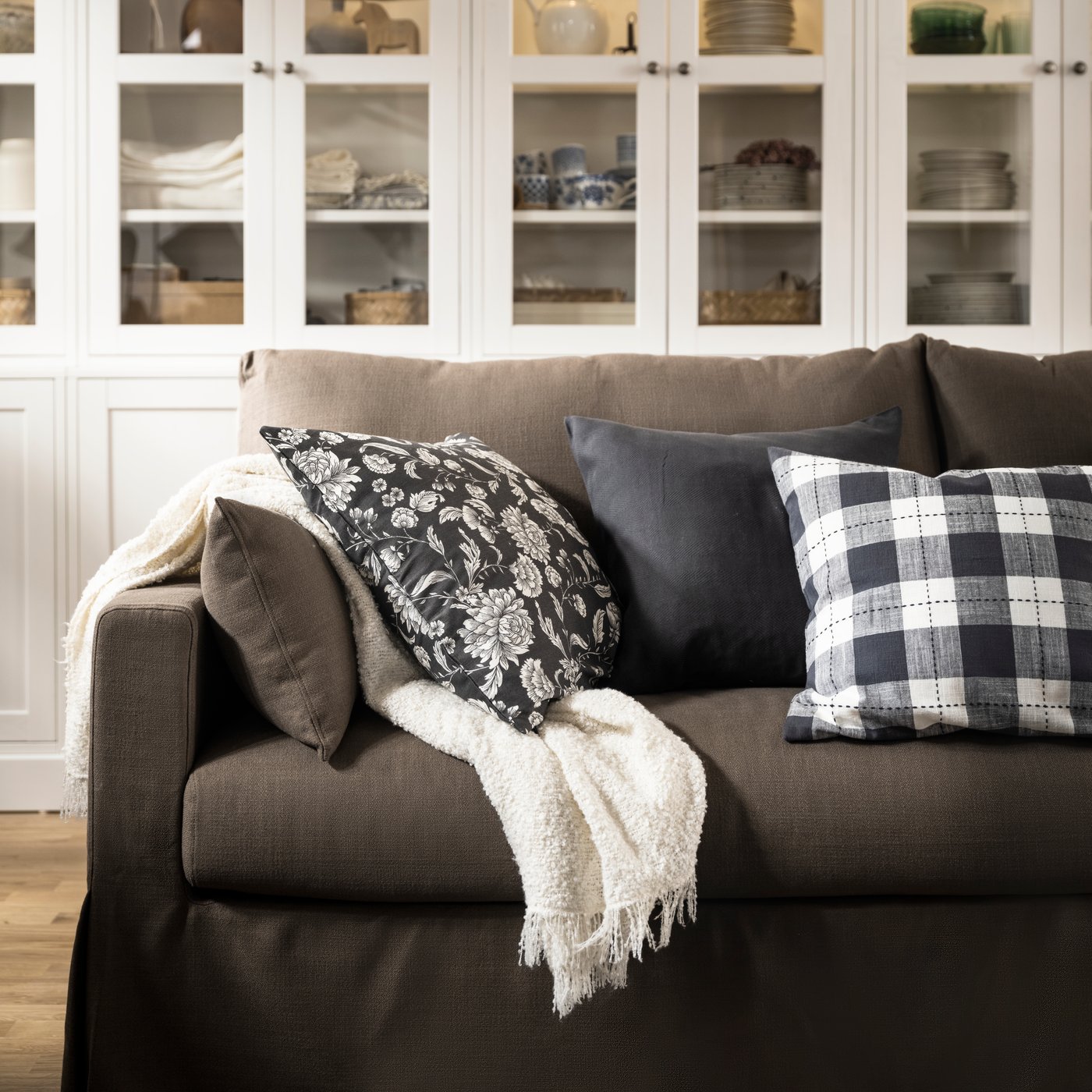 Kremowy pled SANDBRODD i poduszki, w tym jedna w poszewce IDALINNEA z kwiecistym motywem, leżą na sofie HYLTARP.