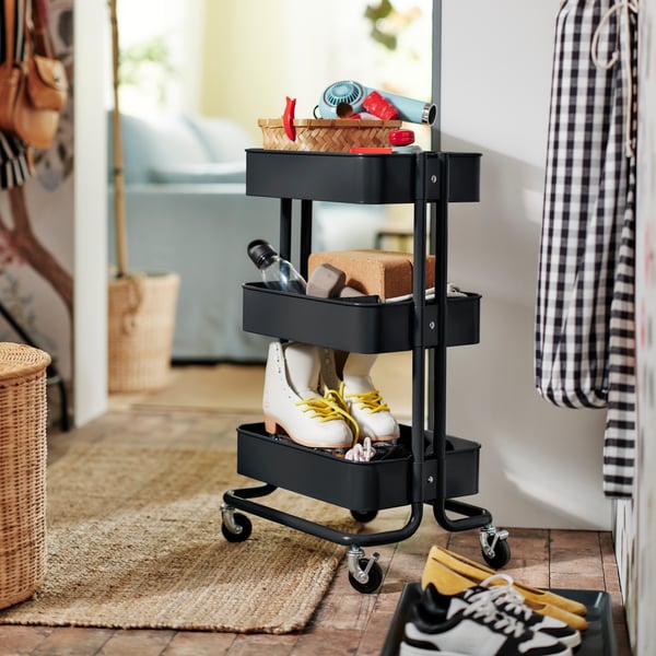 A black RÅSKOG trolley in a hallway on a LOHALS flatwoven rug, holding a hairdryer, exercise gear and rollerskates.
