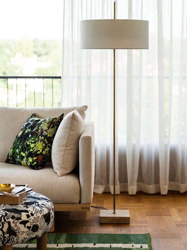 Eine messingbeschichtete weisse Stehlampe STOCKHOLM 2025 auf Parkettboden, vor weißen Fenstervorhängen, mit einem Sofa daneben.