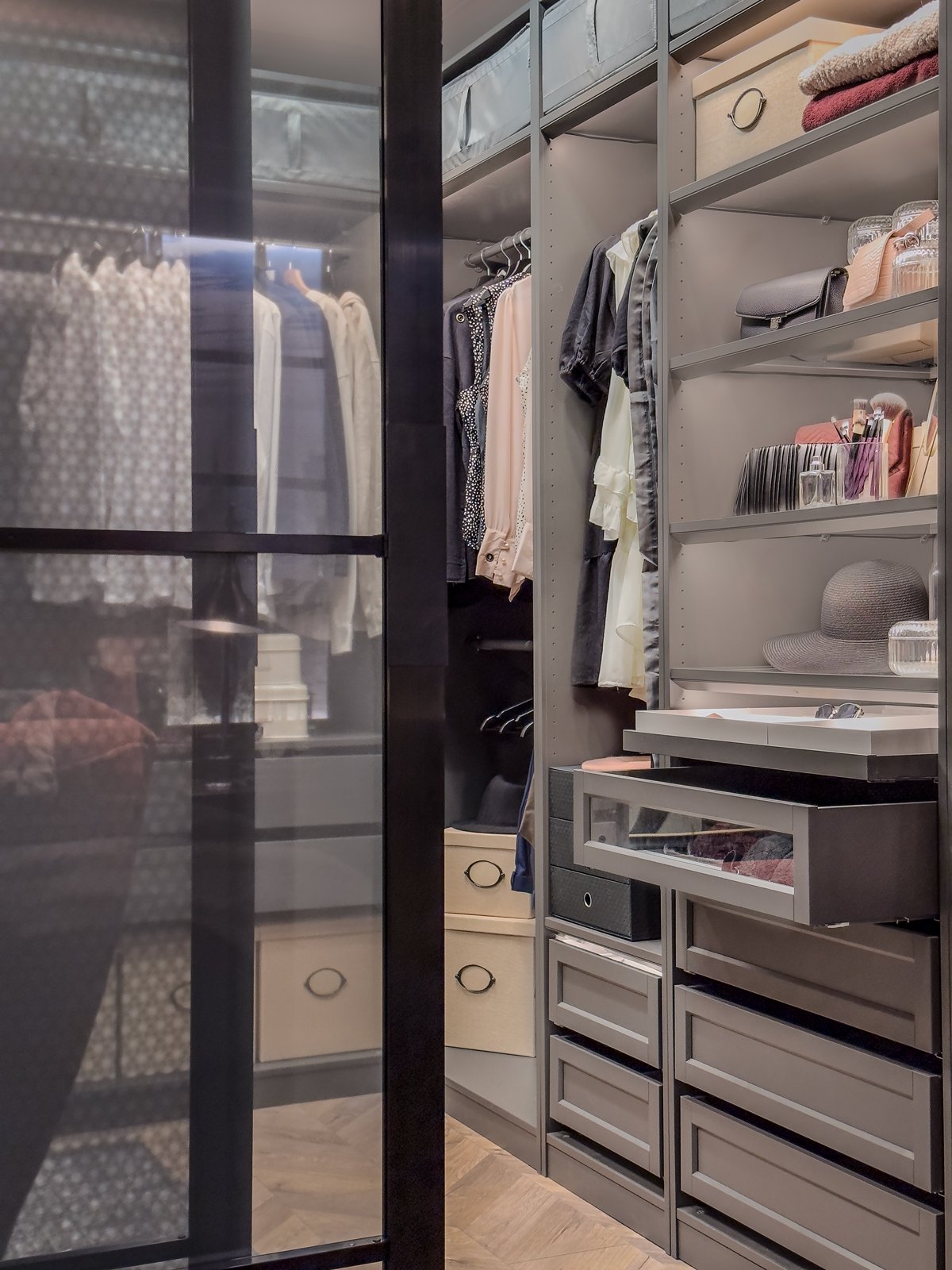 Walk-in closet med öppna hyllor och lådor fyllda med kläder, accessoarer och förvaringslådor, samt skjutdörrar med svart ram och frostat glas.
