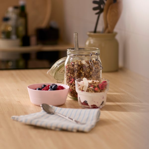 A glass jug with muesli in the back, and two smaller bowls filled with yoghurt and muesli ready to go.