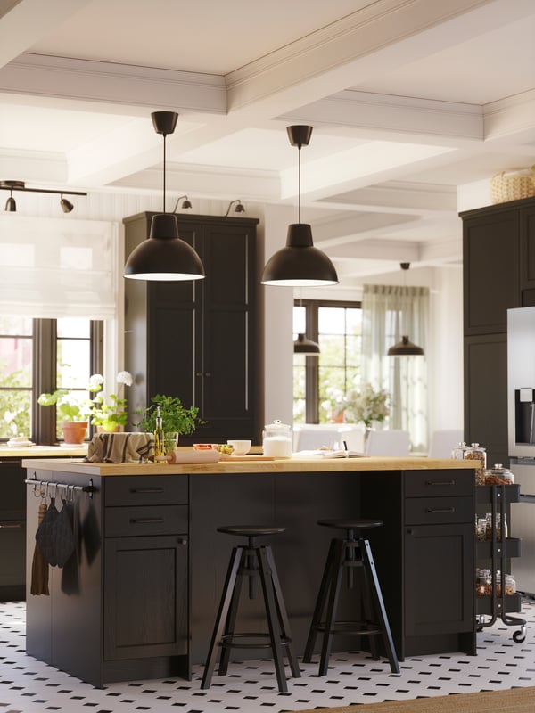 Un îlot de cuisine avec des façades LERHYTTAN teintées noires. Deux tabourets se trouvent devant celui-ci et il est surmonté de deux grandes suspensions noires.
