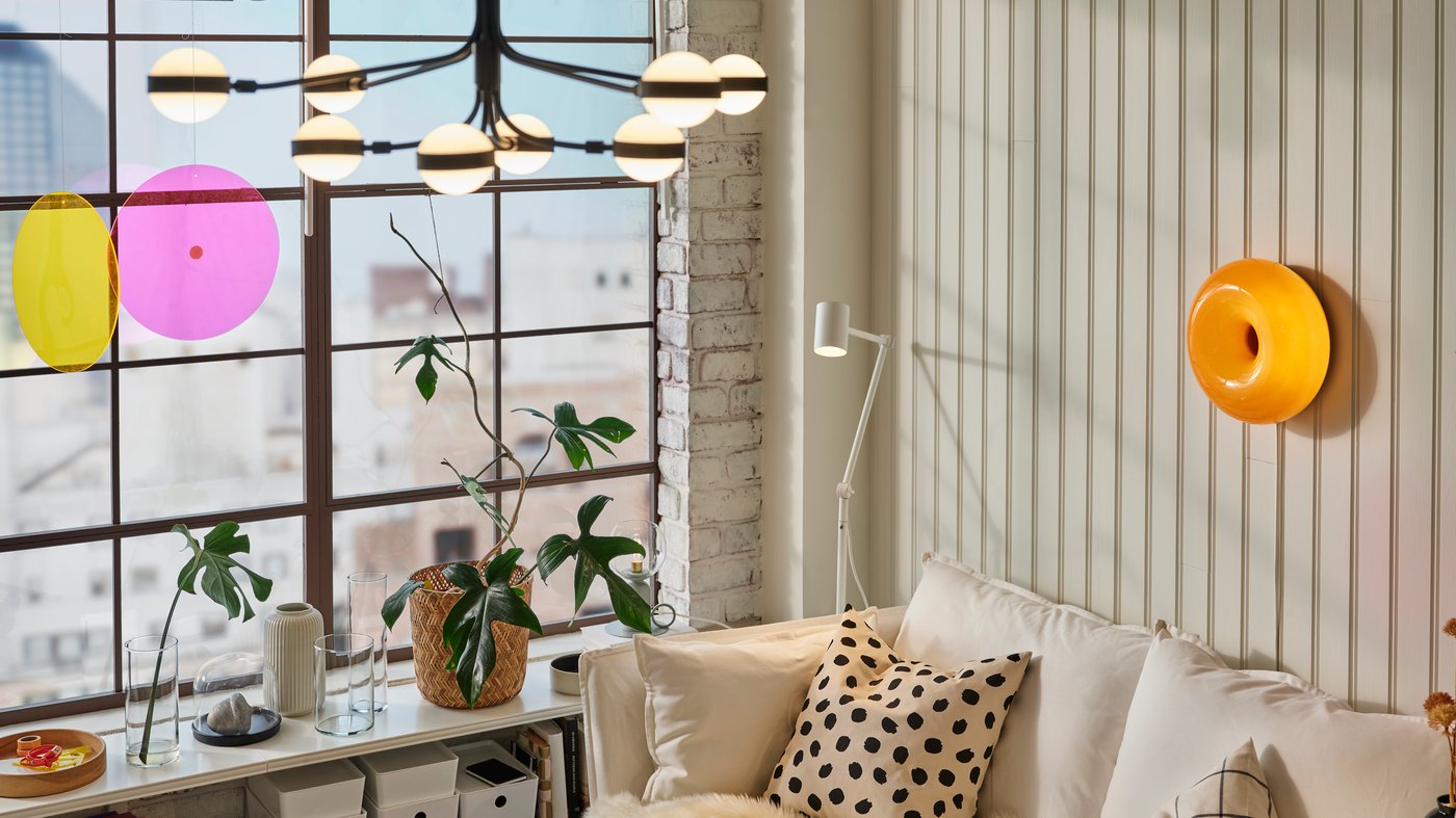 Un lampadario STORSLINGA nero/bianco in una stanza con un divano bianco. Sulla parete lampada da tavolo/parete a LED VARMBLIXT. Accanto al divano si trova una lampada da terra/lettura NYMÅNE bianca e un mobile bianco con sopra dei contenitori con delle piante.