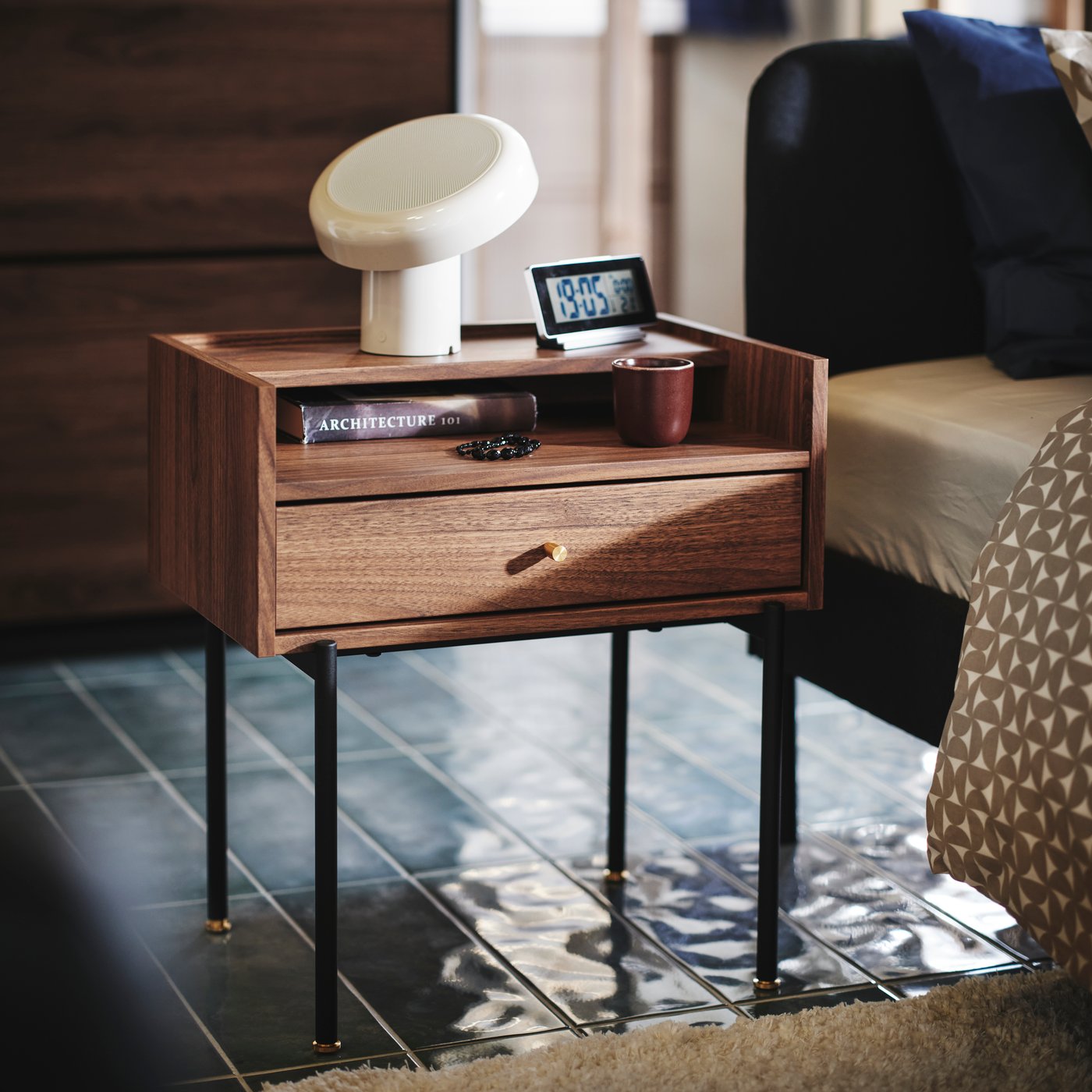 Close-up of RÅDMANSÖ bedside table in a walnut colour with some knick-knacks and BLOMPRAKT Bluetooth speaker lamp on top.