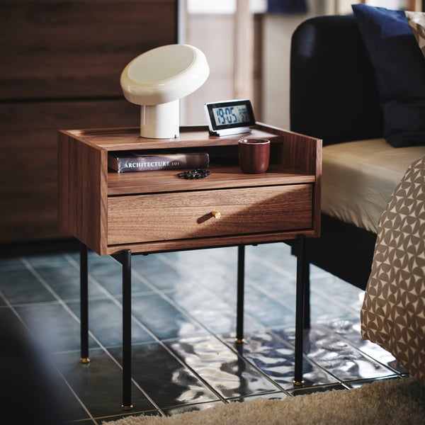 Close-up of RÅDMANSÖ bedside table in a walnut colour with some knick-knacks and BLOMPRAKT Bluetooth speaker lamp on top.